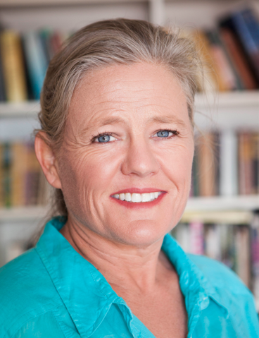 Woman facing camera in turqouise blue shirt, with bookcase blurred as her background